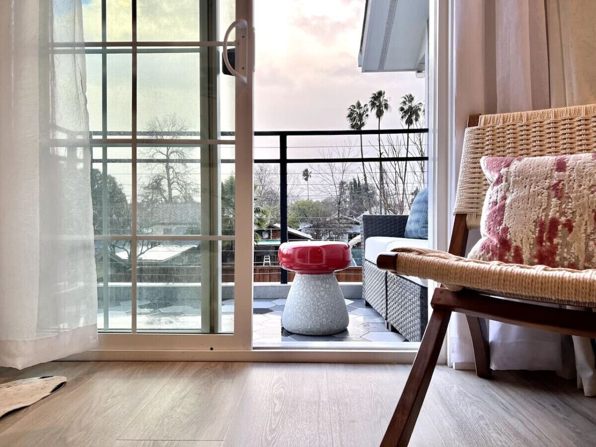 Une pièce dotée d’une porte-fenêtre coulissante donnant sur un espace extérieur avec une terrasse en bois, un fauteuil en osier et un pouf rouge et blanc.