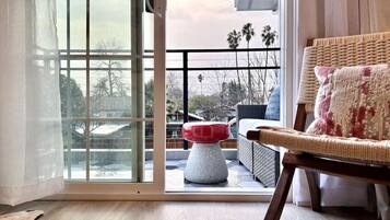 Une pièce dotée d’une porte-fenêtre coulissante donnant sur un espace extérieur avec une terrasse en bois, un fauteuil en osier et un pouf rouge et blanc.