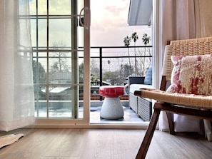 Una habitación con puerta de vidrio corrediza que da a un espacio exterior con terraza de madera, una silla de mimbre y un reposapiés rojo y blanco.