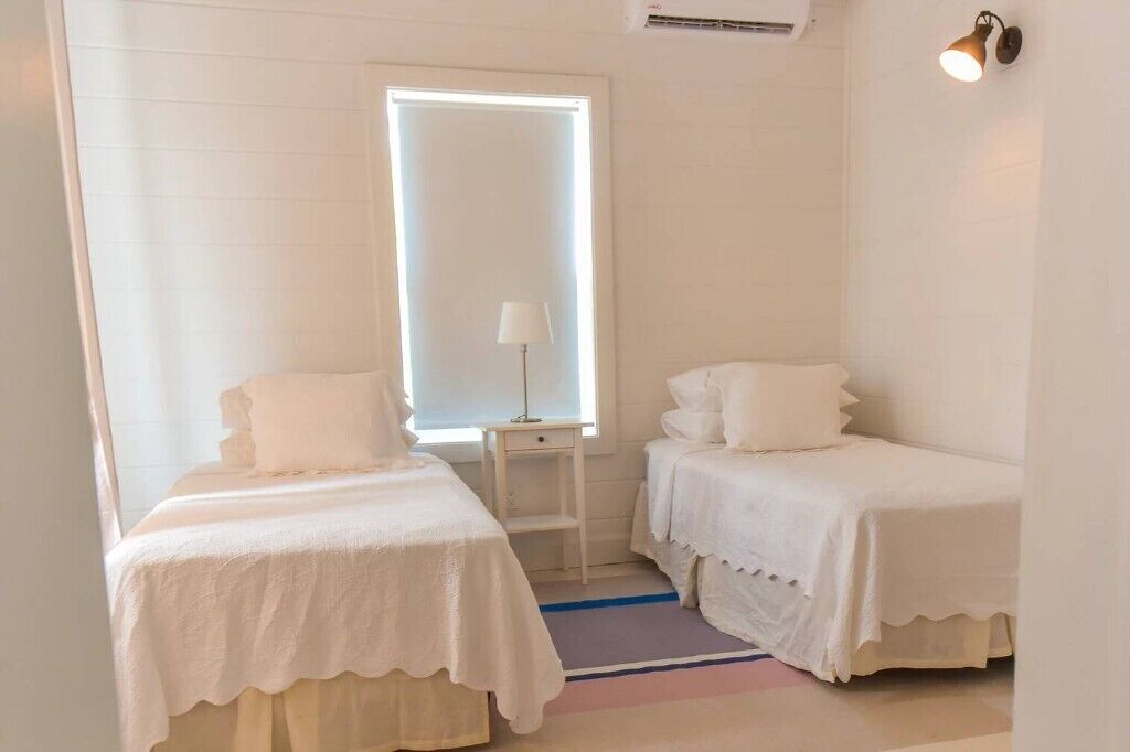 A room with two single beds, a small table, a lamp, and a window with blinds.