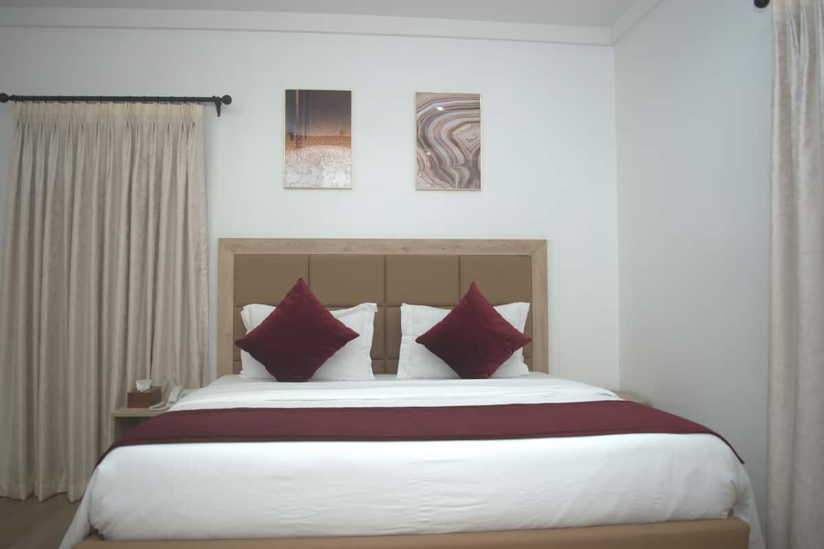 Una cama bien tendida con ropa blanca y dos almohadas rojo oscuro, un cabecero de madera y dos cuadros abstractos en la pared.