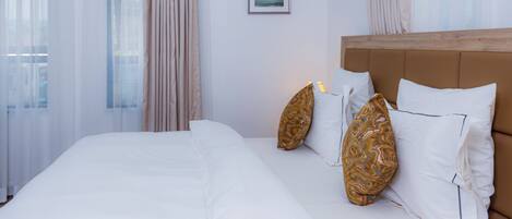 A neatly made bed with white linens and patterned pillows, a wooden headboard, and sheer curtains.
