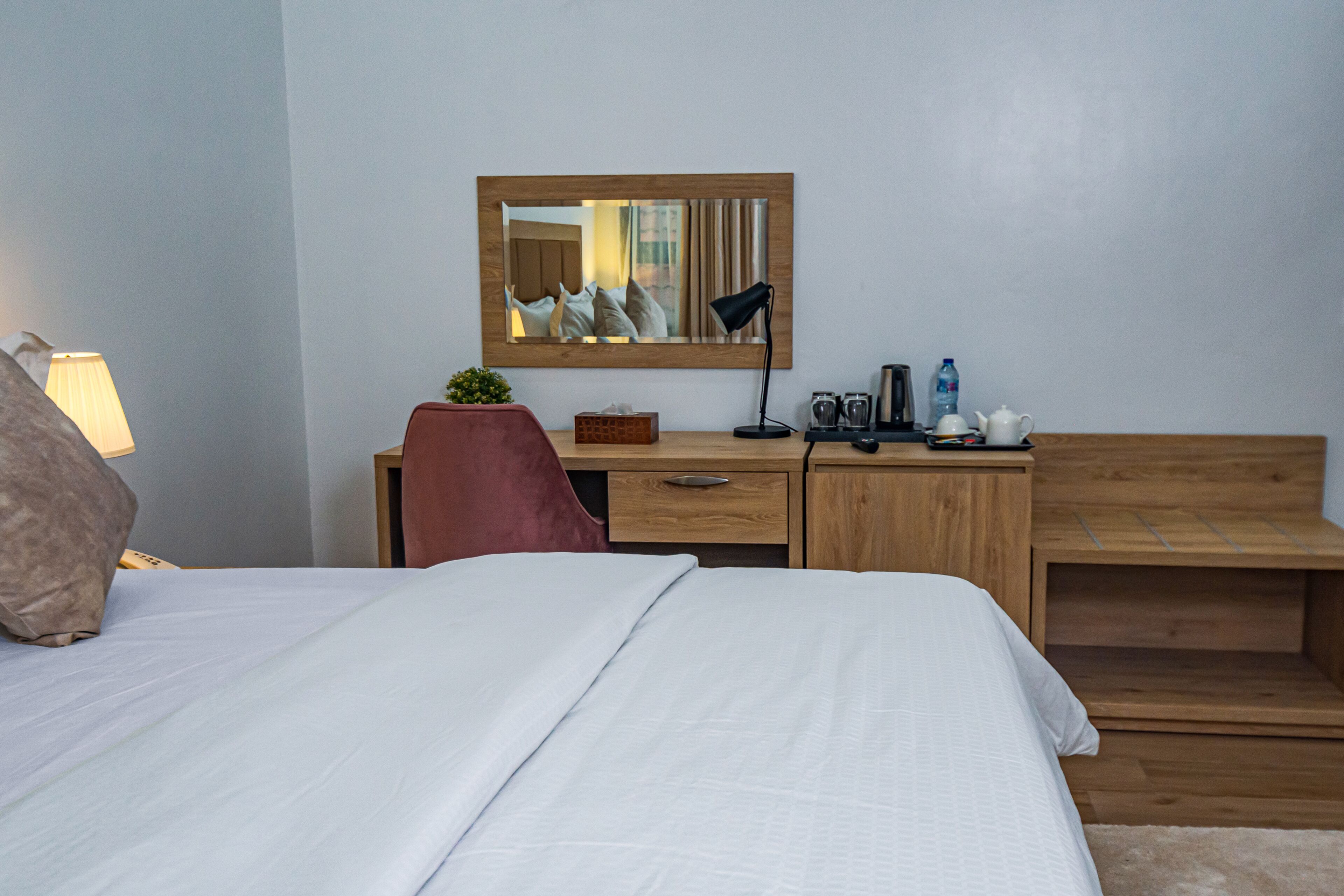 A hotel room with a bed, a desk with a chair, a mirror, a lamp, a small plant, and a wooden bench.