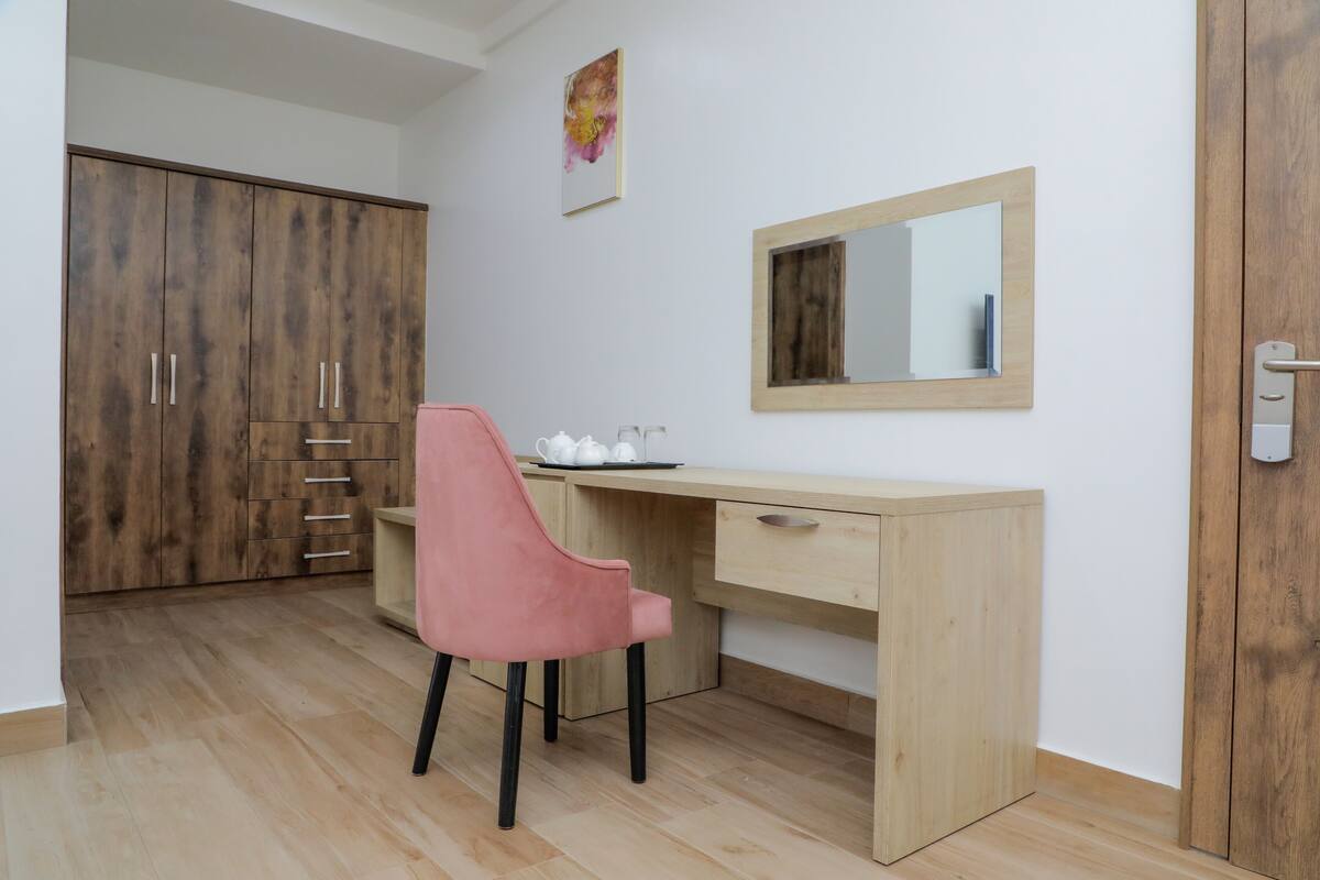 Una habitación con un armario de madera, un escritorio con silla, un espejo y una pequeña mesa con tazas.