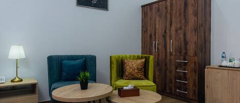 A room with two round tables, a teal sofa, a green armchair, a wooden wardrobe, and a nightstand with a lamp.