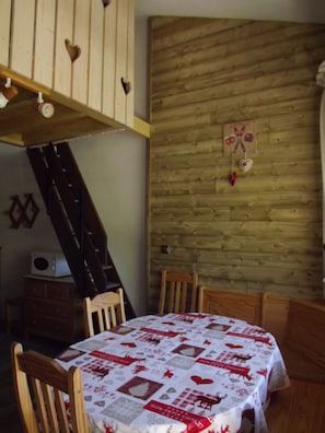 A dining area with a table covered in a patterned tablecloth, wooden chairs, a wooden wall with a heart-shaped decoration, and a staircase with a wooden railing. - Loue aux Menuires (Les Belleville)