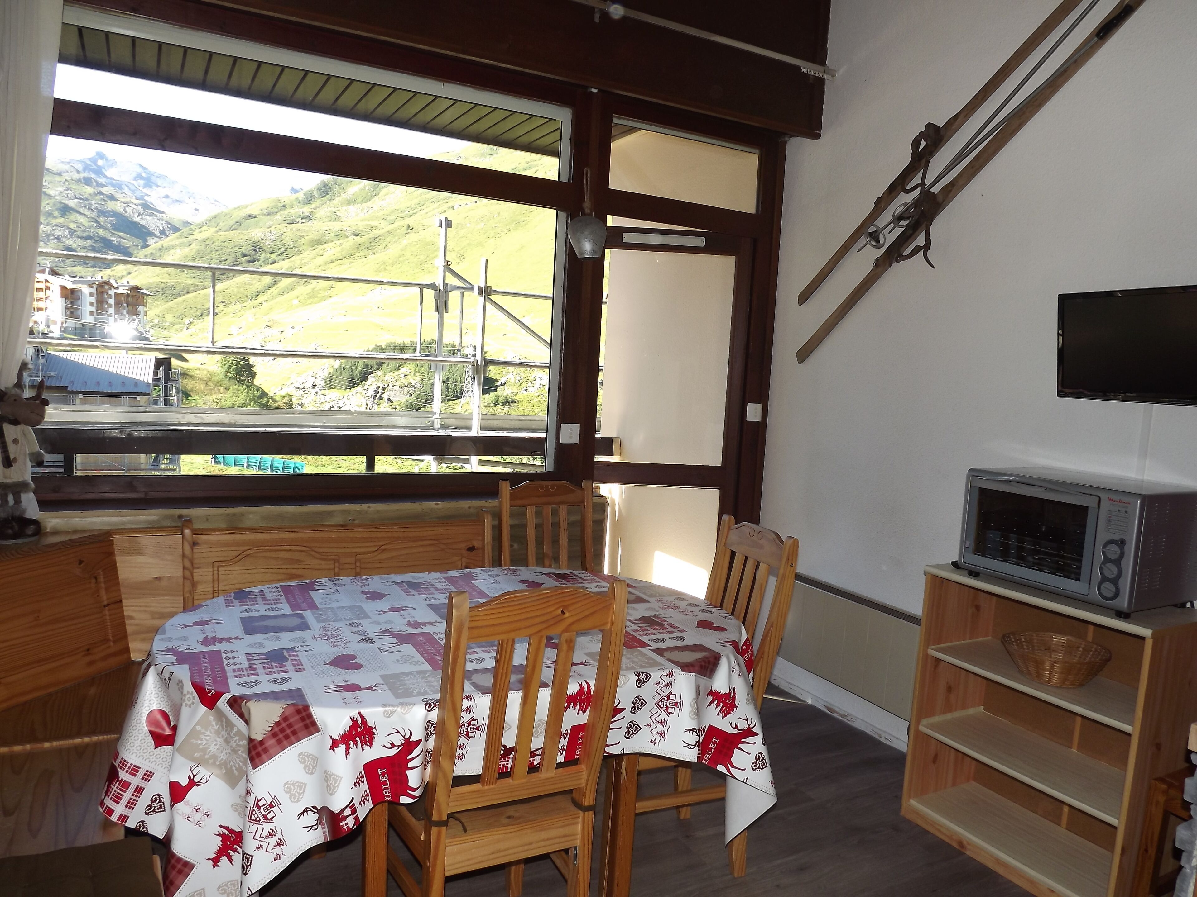 Ein Essbereich mit einem Tisch, der mit einer festlichen Tischdecke bedeckt ist, Holzstühle, eine Holzkommode und ein Regal mit einer Mikrowelle. Ein Fenster bietet Blick auf eine gebirgige Landschaft.