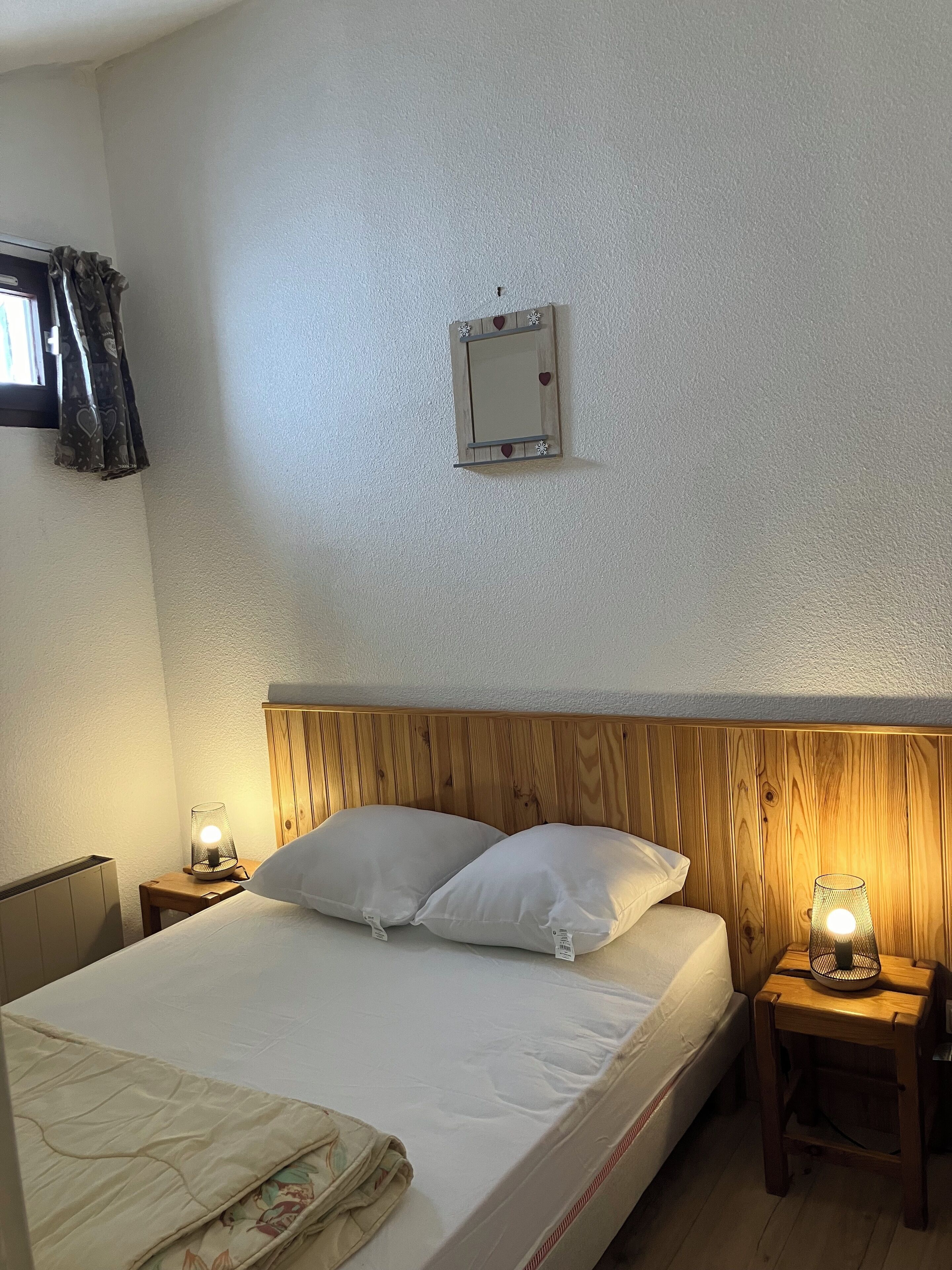 A neatly made bed with white linens, two pillows, and a wooden headboard. There are two bedside tables with lamps, a small wall-mounted light switch, and a window with curtains.
