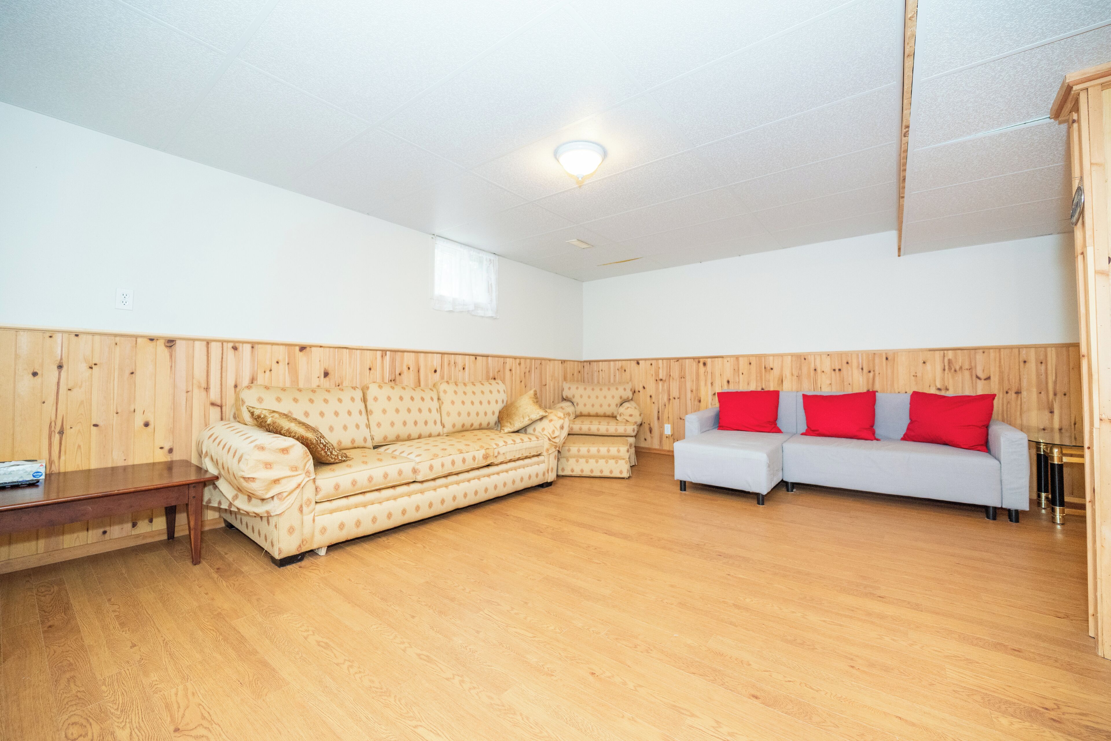 A spacious living room with wooden paneling, a beige sofa, a grey sofa with red cushions, and a wooden coffee table.