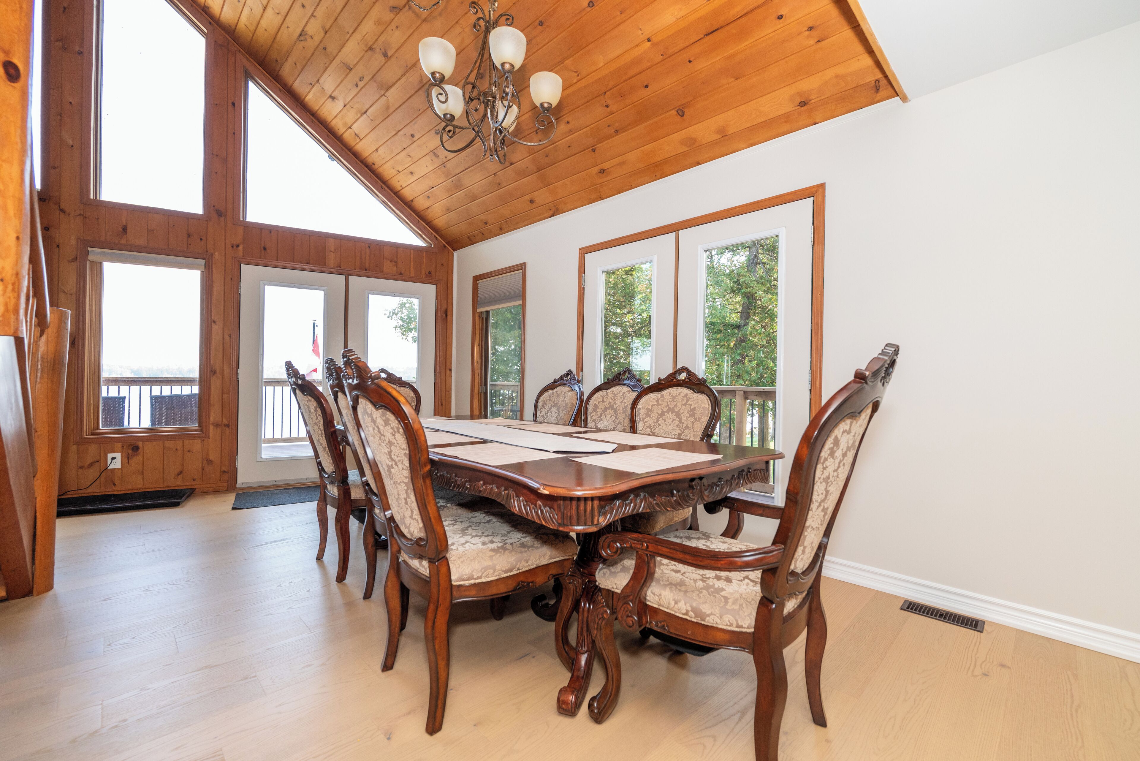 A spacious dining room with a large wooden table and six chairs, high ceilings with wooden beams, and large windows offering views of trees.