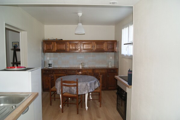 A compact kitchen with wooden cabinets, a dining table with chairs, a white pendant light, and a window with curtains.