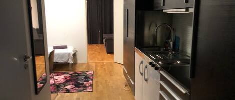 A narrow kitchen with a refrigerator, sink, and cabinets. A small rug and a chair are visible in the background.