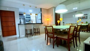 Cocina moderna con armarios blancos, puerta de madera y zona de comedor con mesa blanca y sillas de madera.