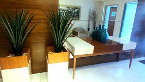 Un área de recepción moderna con un mostrador de madera, dos plantas en macetas y un espejo grande que refleja la estancia.