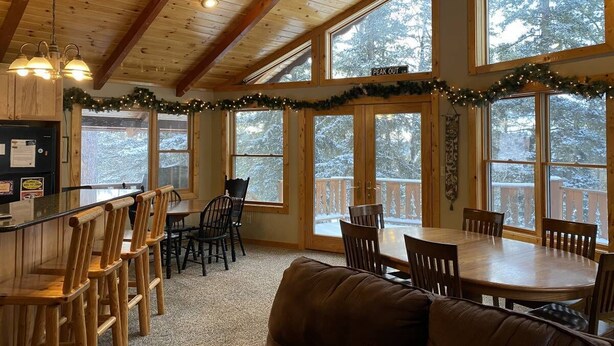 Interior acogedor de una cabaña con comedor, cocina y ventanas amplias que ofrecen vistas a un bosque nevado.