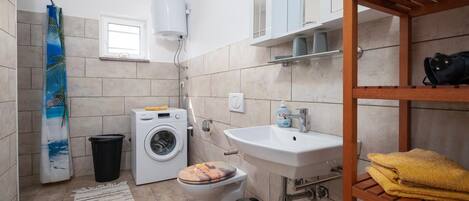 A compact bathroom with a washing machine, toilet, sink, and a wooden shelf with towels.