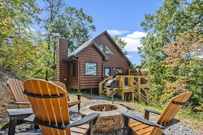 A wooden cabin with a fire pit and Adirondack chairs in front.