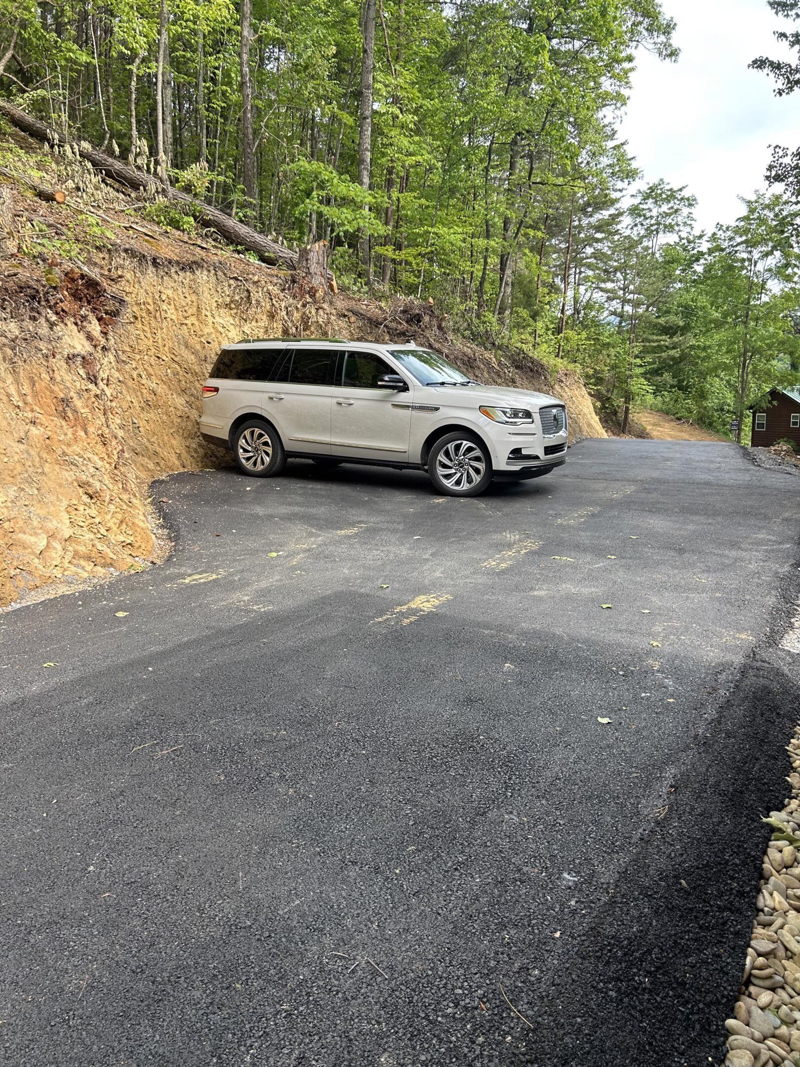 Ein weißer SUV steht auf einer asphaltierten Straße, an einer Seite eine Böschung und im Hintergrund eine Holzhütte.