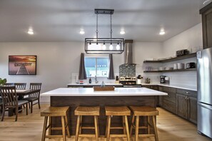 A modern kitchen with a central island, bar stools, a dining area with chairs, and a large window.