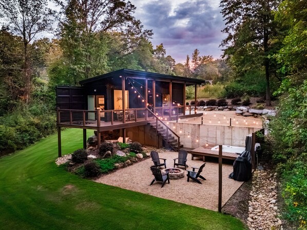 Une terrasse extérieure moderne, dotée d’un foyer et d’un espace salon, entourée d’un jardin aménagé et d’une structure en bois avec un balcon.