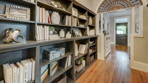 Un ampio corridoio con pavimento in legno, fiancheggiato da librerie piene di libri e oggetti decorativi, che conduce a una porta con un arco decorativo.