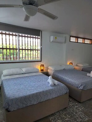 A hotel room with two beds, a ceiling fan, an air conditioning unit, and a window with bars.