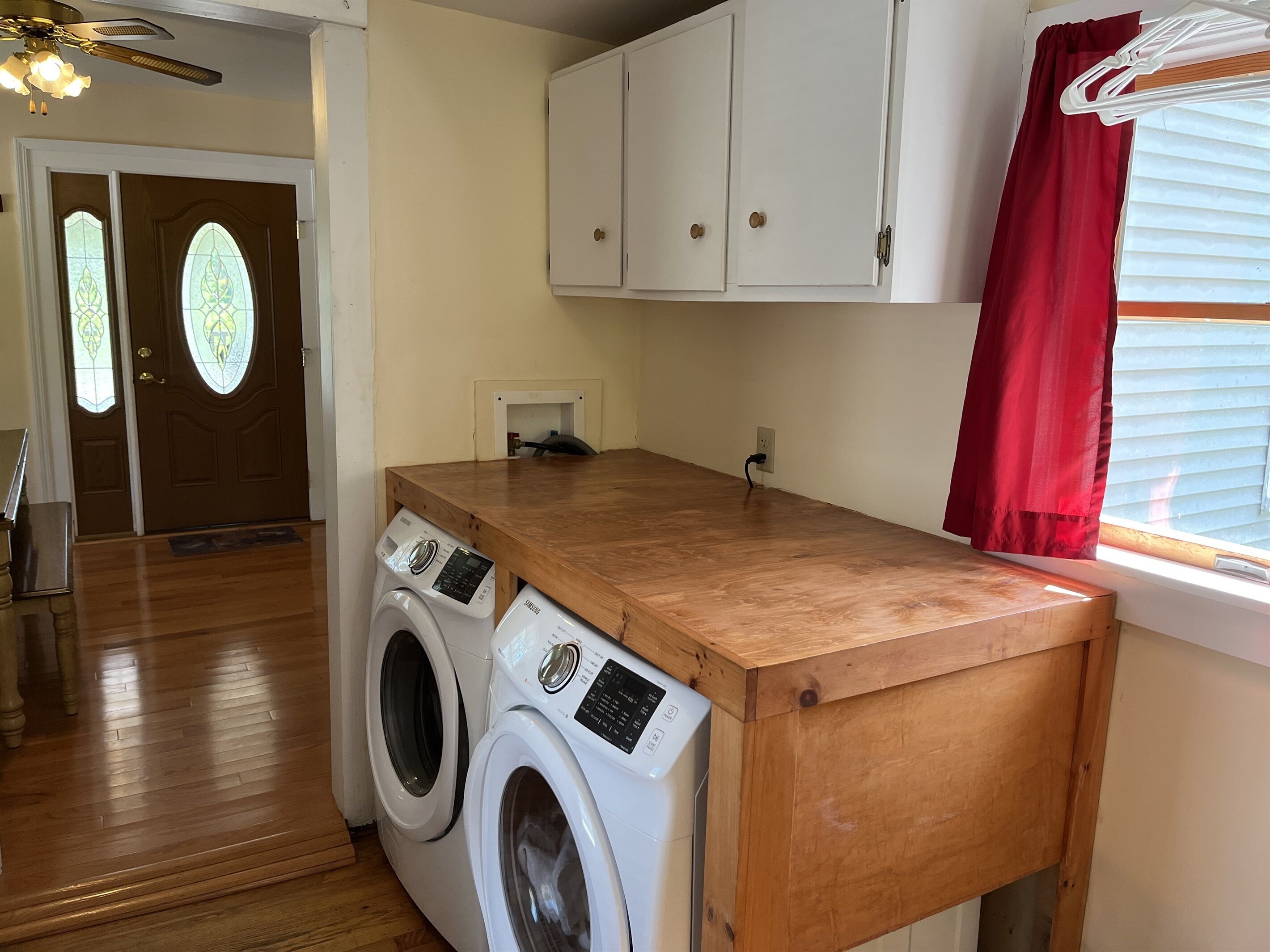 A laundry area with two front-loading washing machines, a wooden countertop, a ceiling fan, and a window with blinds.
