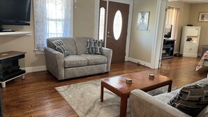 A living room with a grey sofa, a coffee table, a TV on a stand, and a door with a stained glass window. - Alan's Corner - 4 bedroom / 4 bath, pool, less than 5 minutes to Dreams Park (Cooperstown)