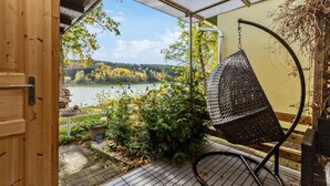 A wooden door leading to a covered outdoor area with a hanging chair, a view of a lake, and some greenery.