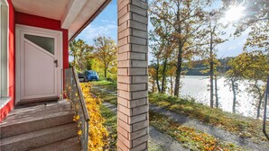 A building with a red exterior, a white door, and a stone pillar. There is a pathway leading to a body of water with trees and a parked car.