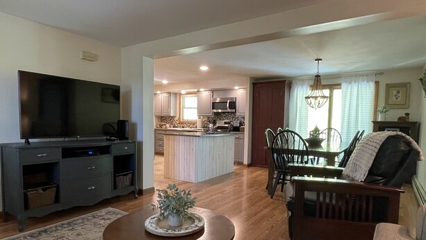 A spacious living room with a large TV, a dining area with a table and chairs, and a kitchen in the background.