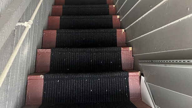 A narrow staircase with black carpeted steps and wooden handrails, leading to a landing area with a window and a hanging light fixture. - Apple Tree Apt 2, Charming 2nd fl apt, under 5 minutes to Dreams Park (Milford)