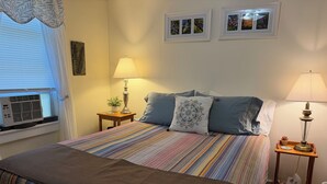 Een bed met een gestreept dekbedovertrek, twee bedlampen, een raam met zonwering en een wandairconditioner.