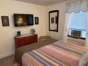A bedroom with a bed, a dresser, a television, a mirror, and a window with an air conditioner.