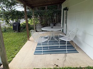 A covered patio area with a round table and four chairs, a trash can, and a white wall.