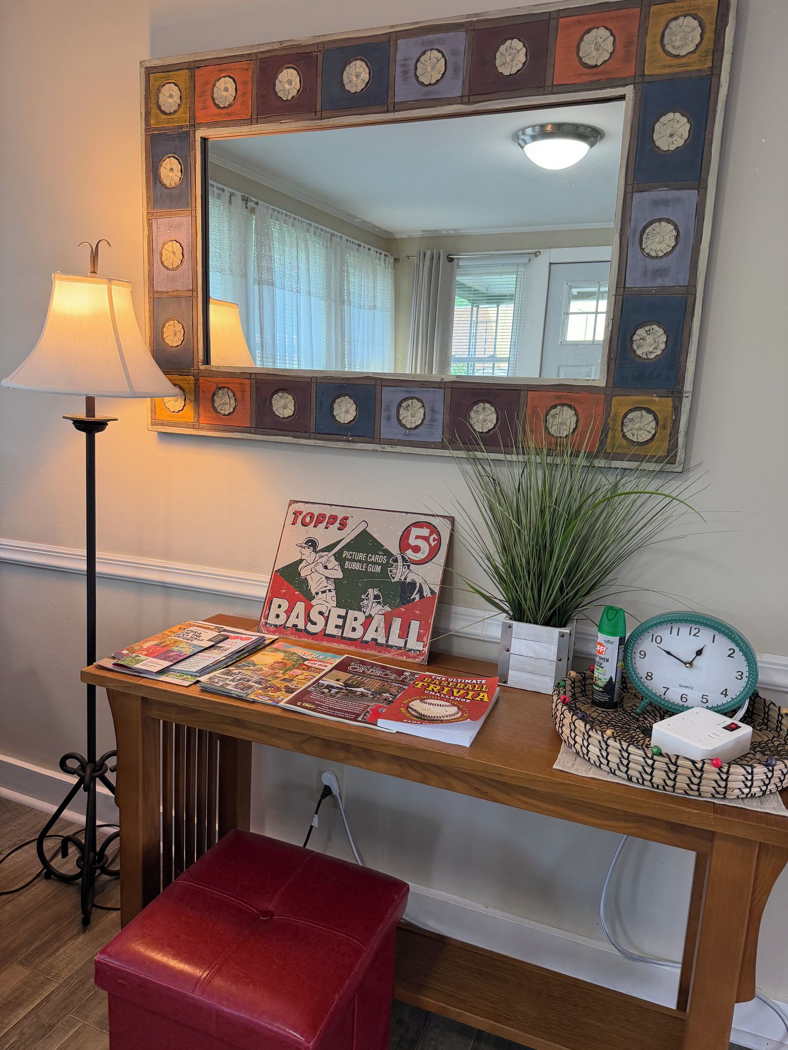 A room with a wooden desk, a mirror with a decorative frame, a lamp, a plant, a clock, and a red ottoman.