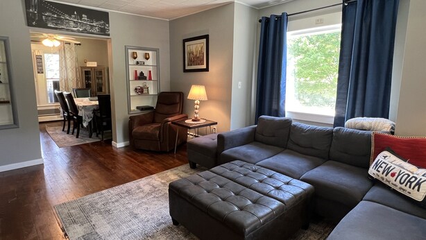 Uma sala de estar com sofá seccional, uma poltrona de couro marrom, uma área de jantar com mesa e cadeiras, um abajur e uma janela com cortinas azuis.