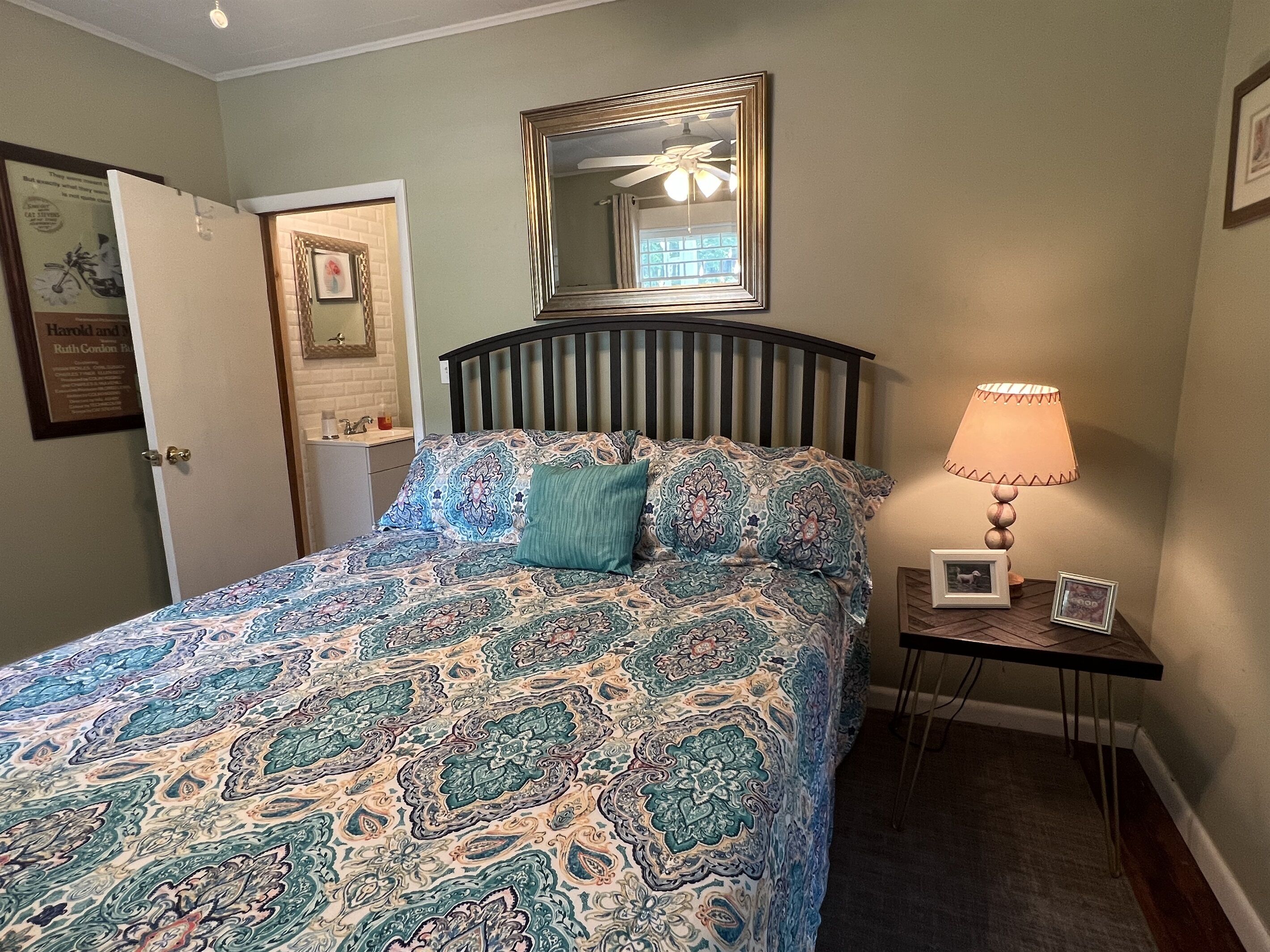 A bedroom with a patterned bedspread, a bedside lamp, a framed picture, and a mirror on the wall.
