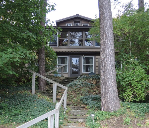 A two-story house with a wooden staircase leading to the entrance, surrounded by dense greenery and trees.