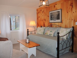 A cozy bedroom with a wooden wall, a bed with striped bedding, a bedside table with a lamp, and a window with curtains.