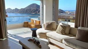 A spacious living room with a large window offering a view of the sea and mountains. There is a sofa with cushions and a coffee table with decorative items.