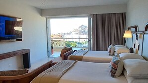 A hotel room with two beds, a desk with a computer, and a large window offering a view of the city.