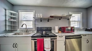 Una cocina con estufa de acero inoxidable, microondas, lavavajillas y un lavabo. Hay dos ventanas con persianas, un estante con vajilla y una toalla colgada en la puerta del horno.