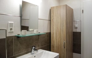 A bathroom with a white sink, a mirror, a glass shelf with toiletries, a wooden cabinet, and a shower area. - Beautiful home in Heckenmünster (Heckenmünster)