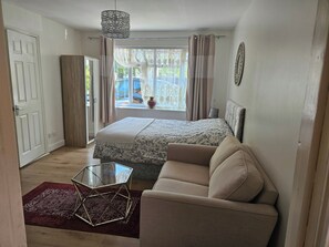 Un dormitorio con cama, sofá, mesa de vidrio, alfombra roja, ventana con cortinas y lámpara colgante.