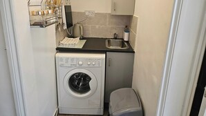 A compact kitchen with a washing machine, a small sink, and a countertop with a kettle and utensils. - Lovely Studio in Town Centre (Reading)