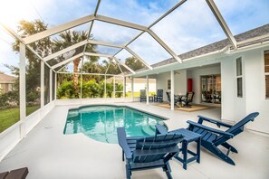 A covered swimming pool with lounge chairs and a table, surrounded by a white exterior wall and glass doors.