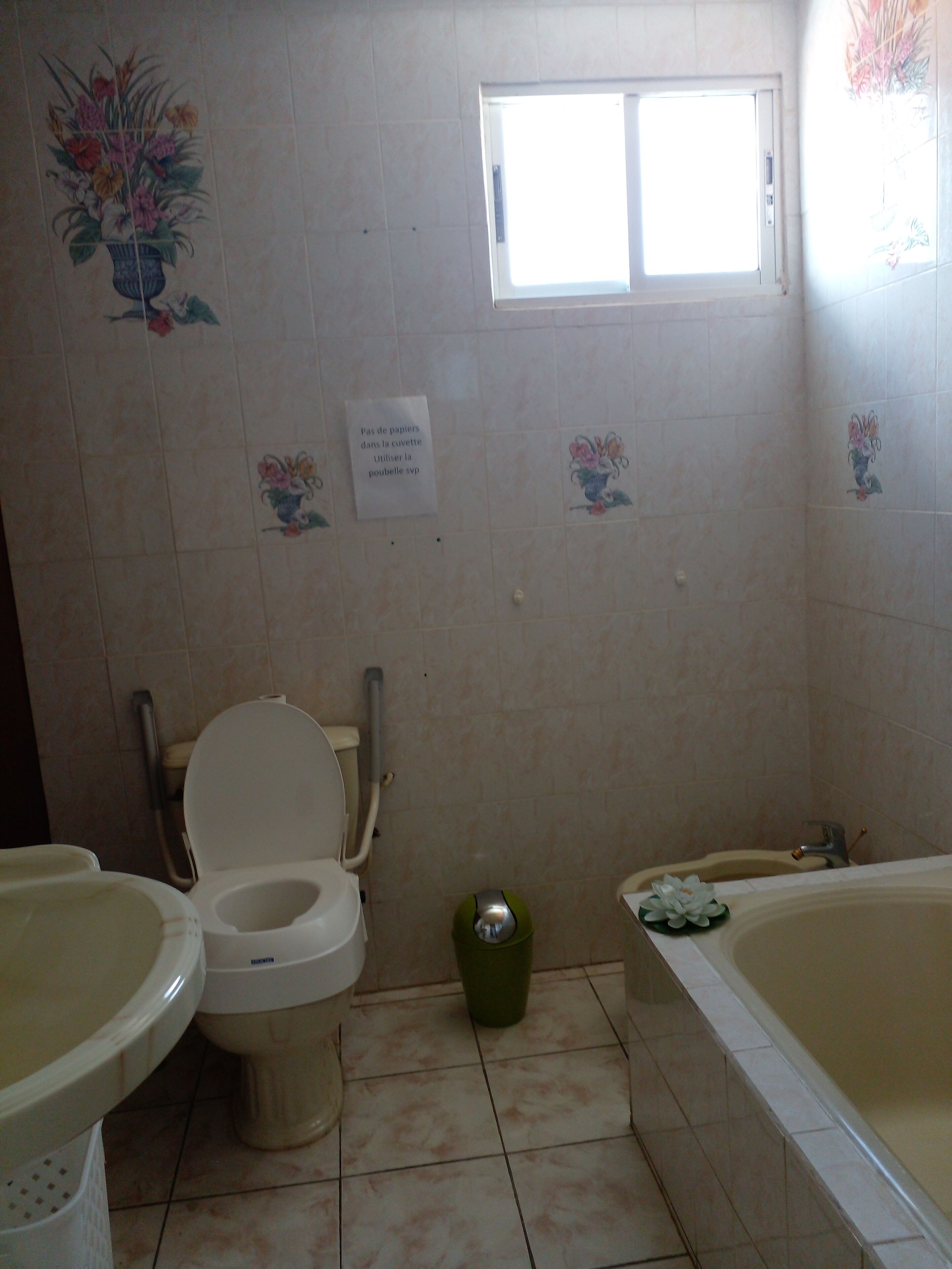 A bathroom with a toilet, bidet, and bathtub. There is a window above the toilet and a sign on the wall.