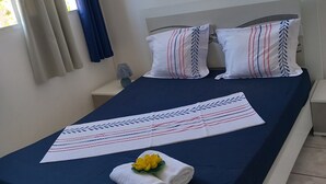 Una cama bien tendida con una colcha azul, almohadas blancas con rayas rojas y azules, y una toalla blanca con una flor amarilla.