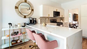 Een moderne keuken met witte kasten, een centraal eiland, roze barstoelen en een ronde spiegel aan de muur.